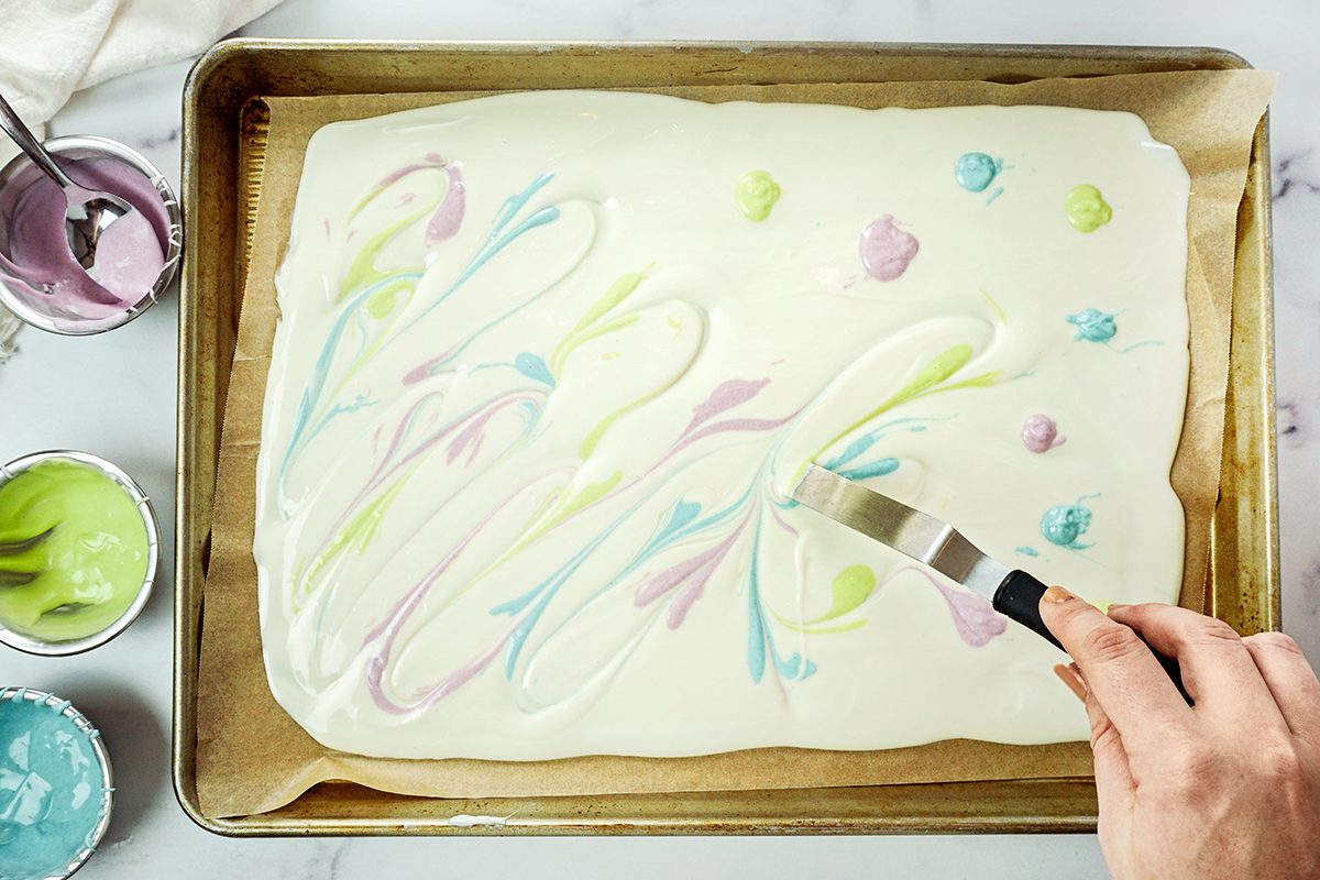 A hand uses a spatula to create swirls of pastel colors like pink, purple, blue, and green on a layer of white frosting on a rectangular cake. Small bowls of colored frosting are nearby on a marble surface.