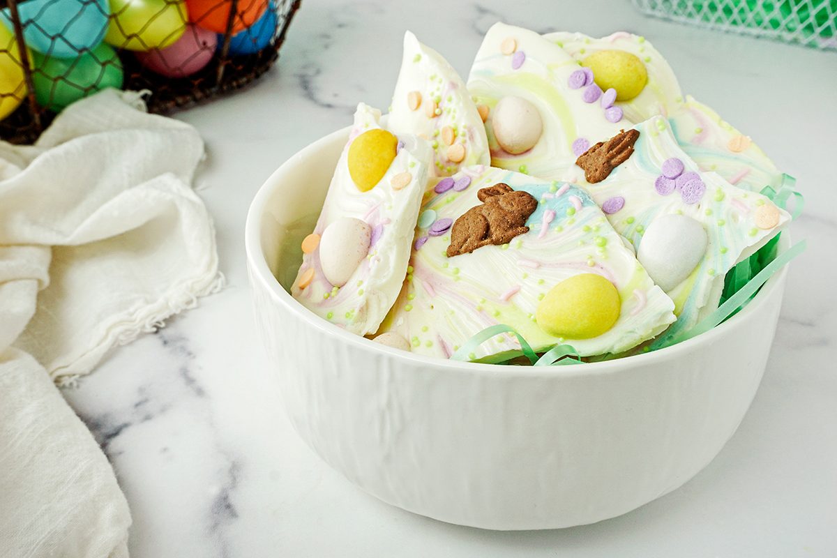 A white bowl filled with colorful Easter-themed bark featuring pastel candies, chocolate pieces, and various small decorative elements. A basket with colorful eggs is partially visible in the background on a marble surface.