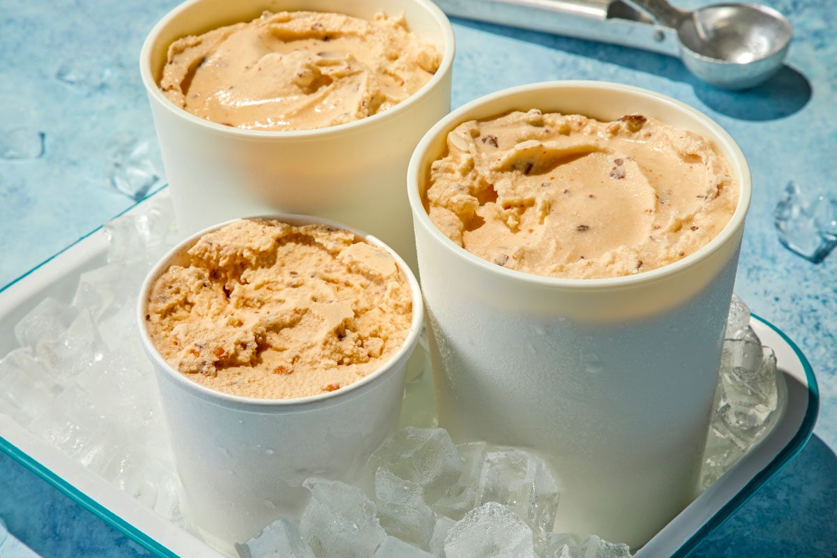 3/4th shot of three containers of Butterfinger Ice Cream, positioned within a bed of ice, Each container is filled with a generous scoop of ice cream, The background features a soft, light blue color