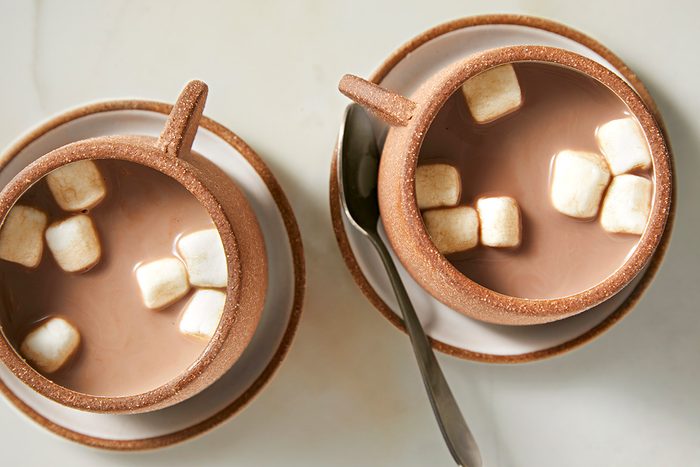 Two cups of hot chocolate topped with marshmallows sit on saucers. A spoon rests on one saucer. The cups and saucers are a matching light brown color, and the scene is set on a light-colored surface.