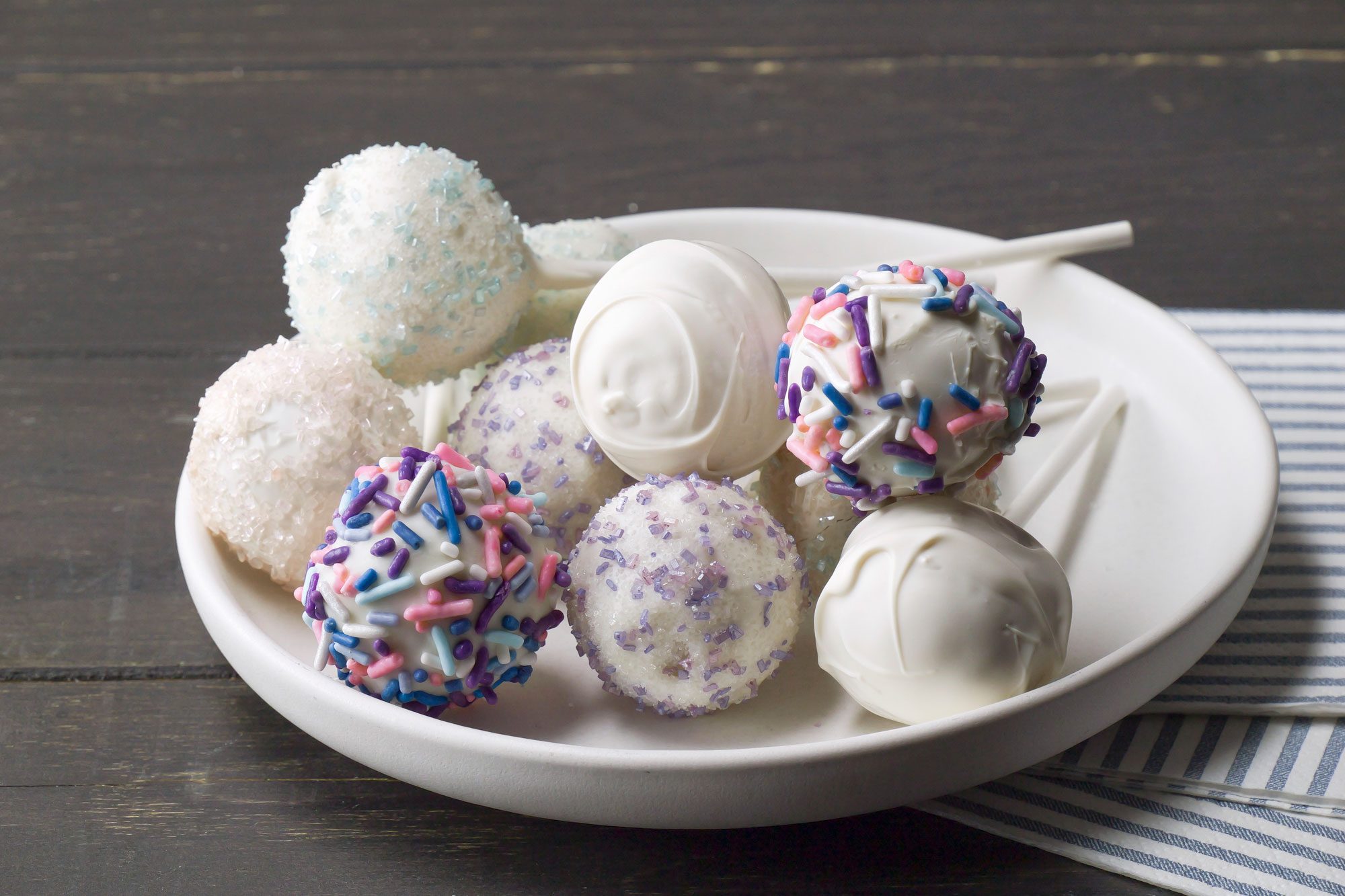 closeup shot of of cake pops; the pops are round and coated in various decorative toppings, they are presented on a simple white plate, in the background, a striped blue and white napkin