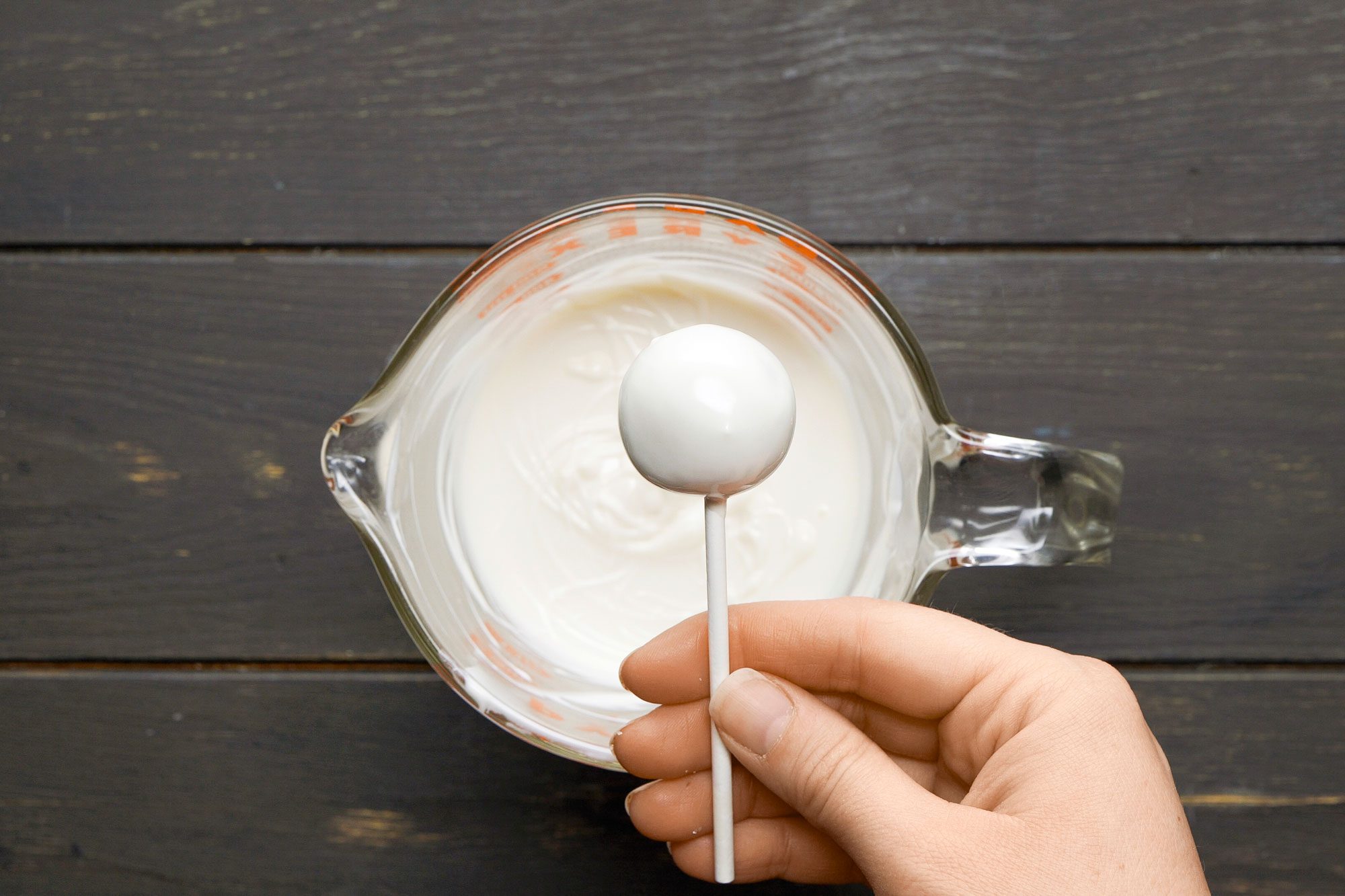 overhead shot of a hand holding a cake pop; the background includes a clear glass bowl filled with a creamy white mixture, the surface beneath is dark wood