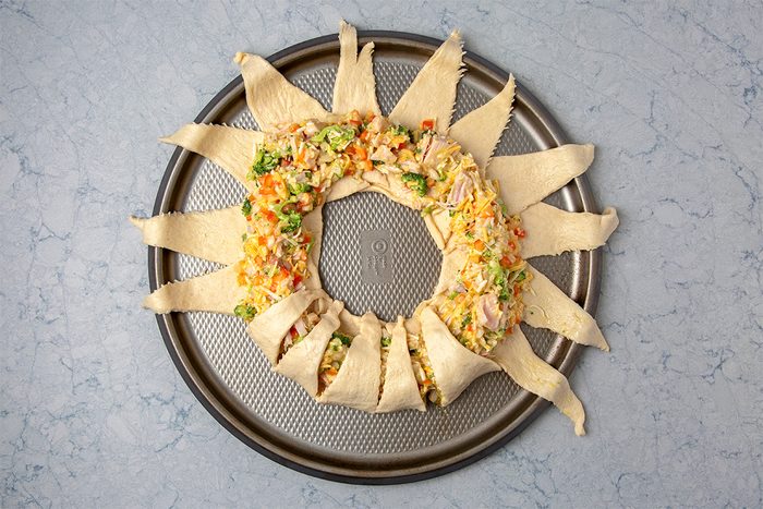A circular arrangement of crescent roll dough on a baking sheet, filled with a mixture of diced vegetables and meat, resembling a wreath. The dough is folded over the filling, ready to bake.