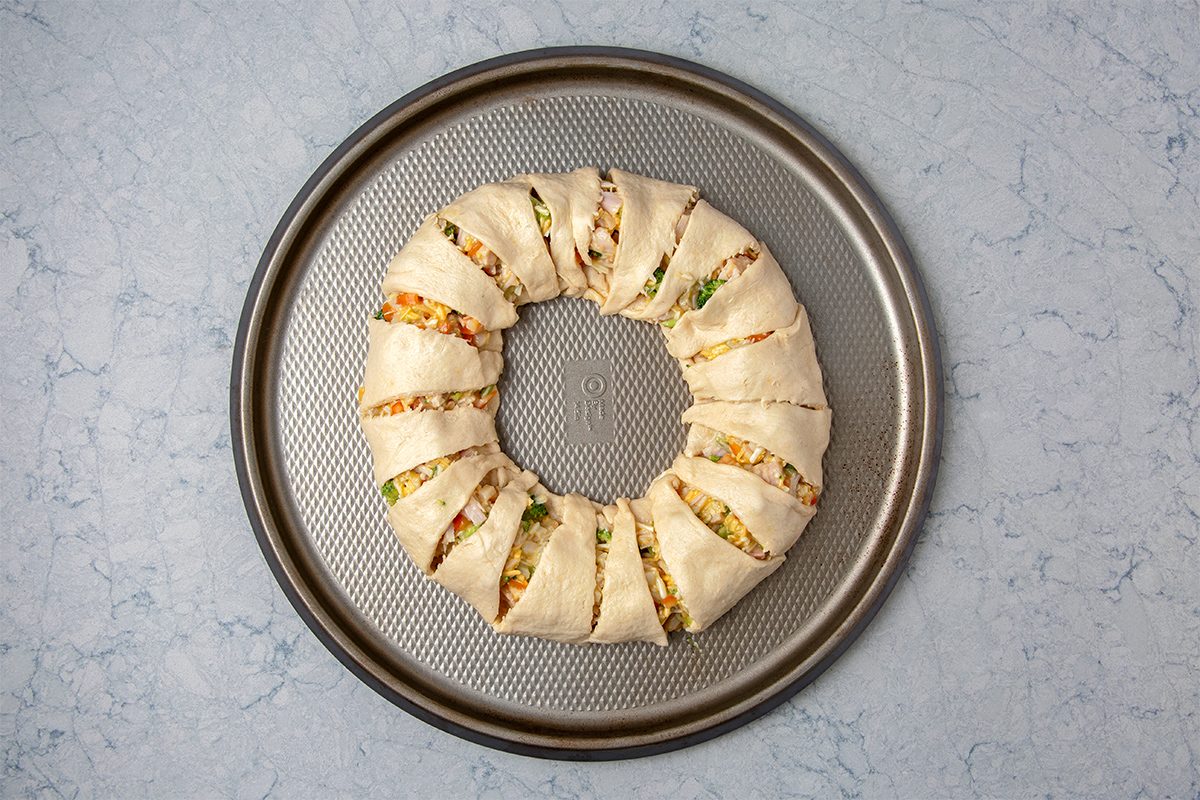 A round, unbaked, stuffed pastry wreath is on a circular baking sheet. The dough is folded over filling that includes vegetables and cheese, arranged in segments, ready for baking. The background is a light marbled surface.