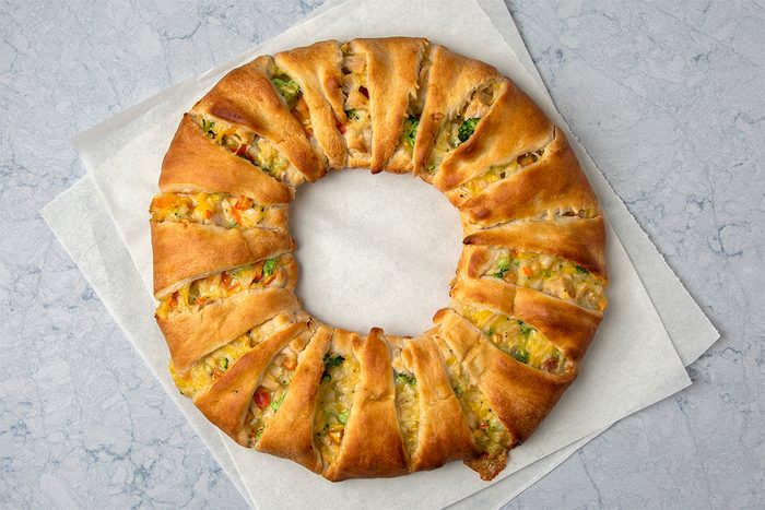 A round, golden-brown baked ring sits on a light-colored surface. The bread is sliced and filled with a mixture of colorful vegetables and cheese, creating a visually appealing pattern. The backdrop is plain and minimalistic.