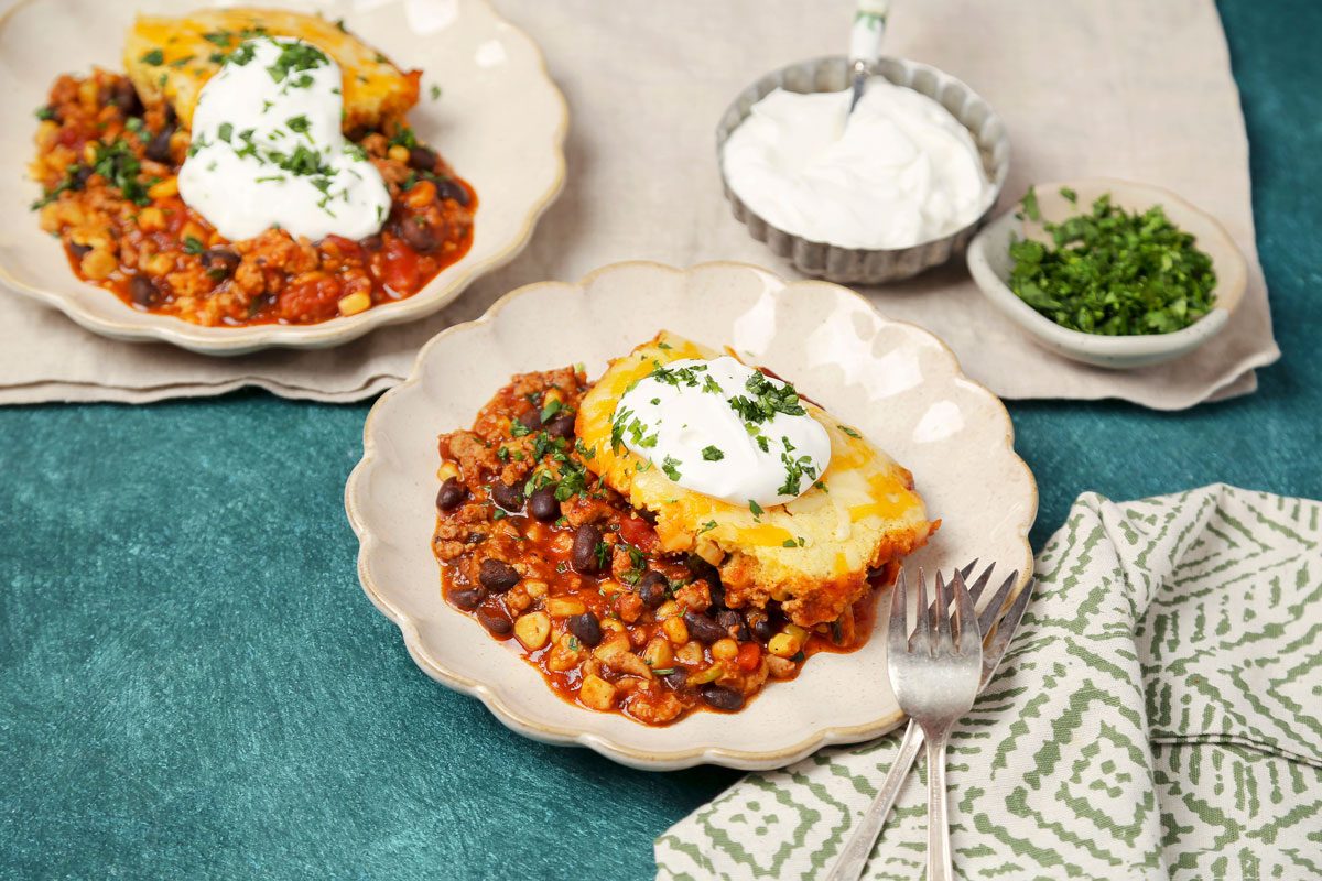 Table view shot of Easy Chicken Tamale Pie; cut and serve on plates; sour cream on top; sprinkle chopped cilatnro; forks; napkin; green surface;