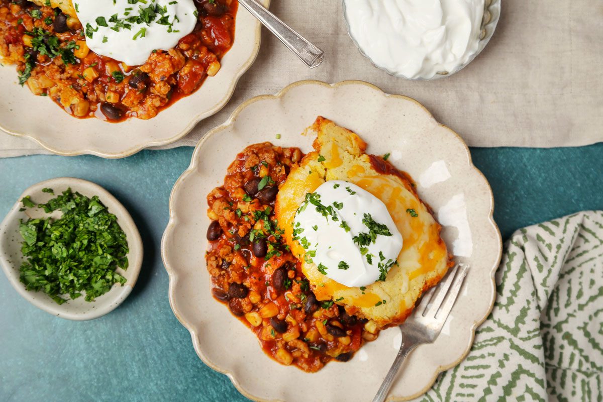 Overhead shot of Easy Chicken Tamale Pie; cut and serve on plates; sour cream on top; sprinkle chopped cilatnro; forks; napkin; green surface;