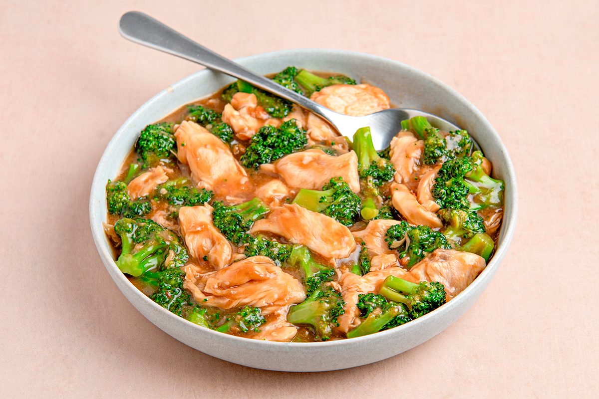 A bowl of chicken and broccoli stir-fry in a savory sauce, served with a spoon on a beige background. The dish features tender chicken pieces and vibrant green broccoli florets.