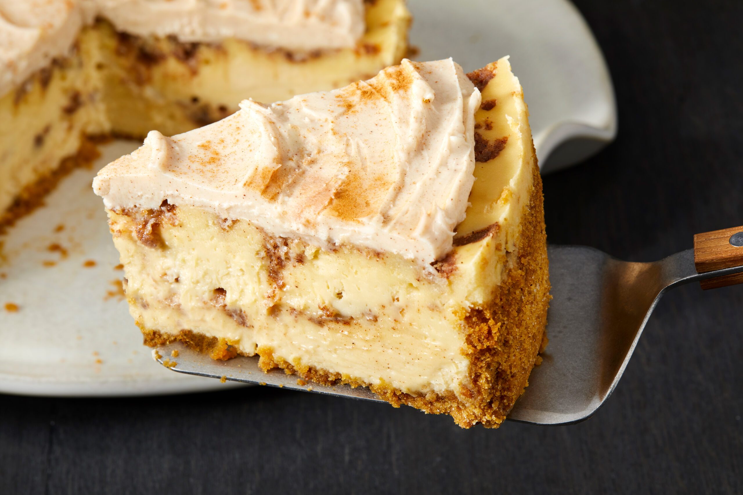 A slice of cinnamon roll cheesecake with a creamy topping and cinnamon swirls on a spatula. The cheesecake has a golden-brown crust and is placed on a white plate. The background is a dark surface.