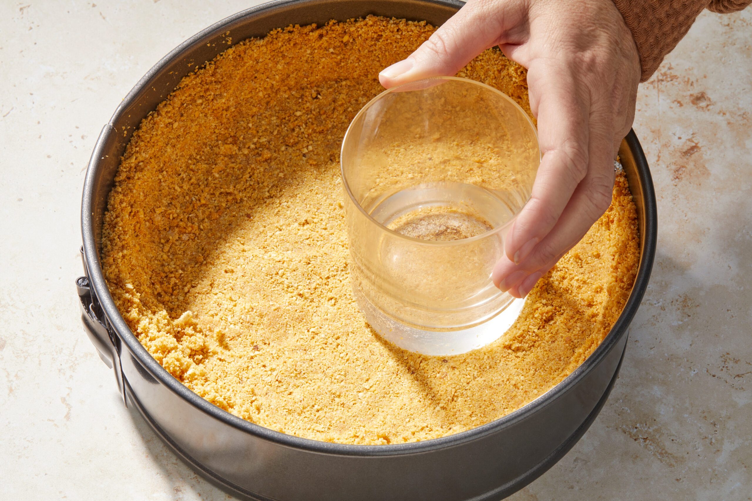 A person presses a flat-bottomed glass onto a crumbly crust in a round springform pan. The crust is made of finely ground crumbs and the person's hand is visible, smoothing the crust surface. The background is a light, textured surface.