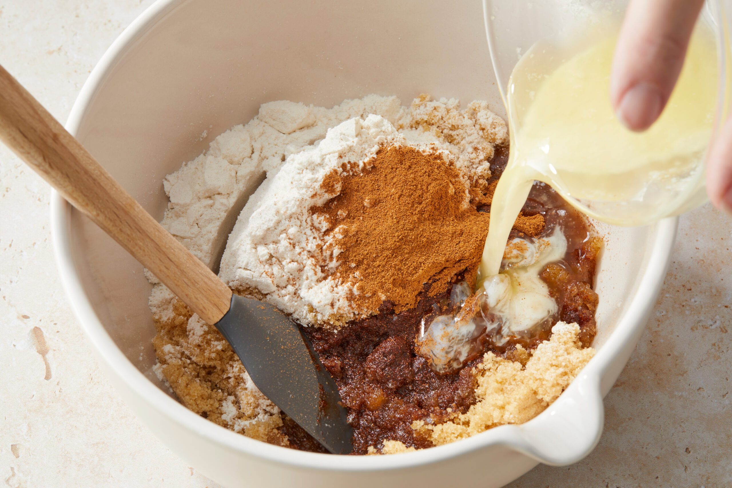 A white bowl contains flour, brown sugar, cinnamon, and other ingredients being mixed. Liquid is being poured into the bowl, and a spatula is resting inside. The preparation appears to be for baking.