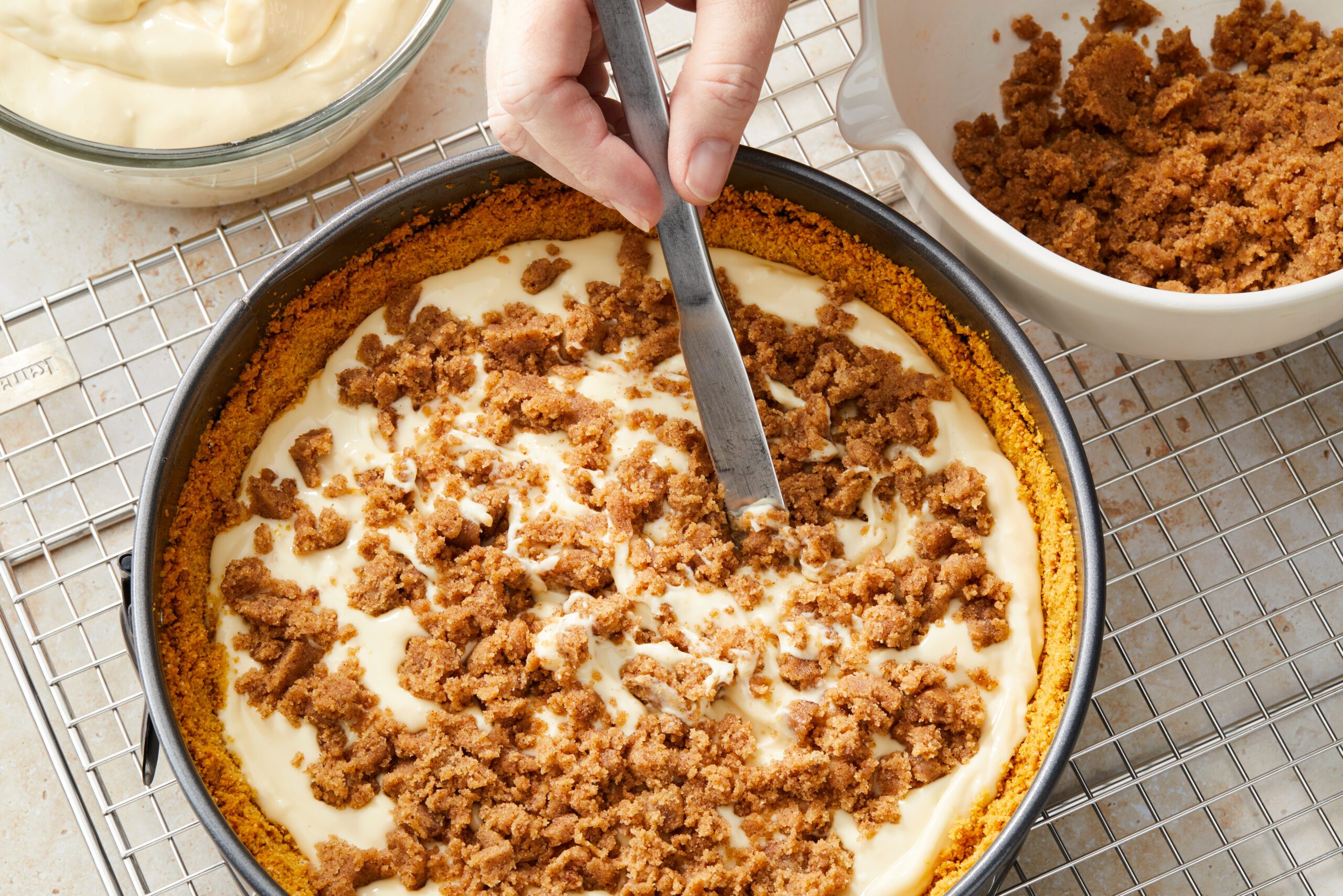 A hand uses a flat utensil to smooth cream over a cheesecake topped with crumbly cookie pieces in a round pan. The pan sits on a cooling rack with bowls of cream and cookie crumbs nearby.