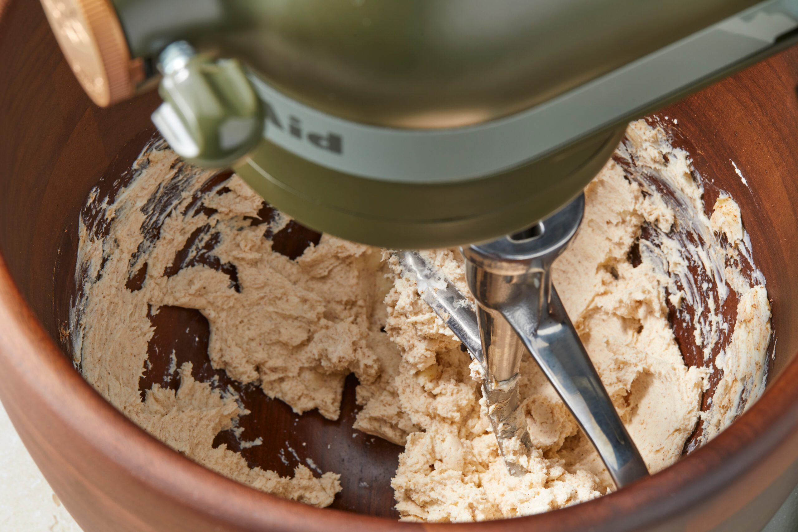 A green stand mixer is shown mixing dough in a brown bowl. The dough is partially blended, and the metal mixing attachment is visible inside the bowl.