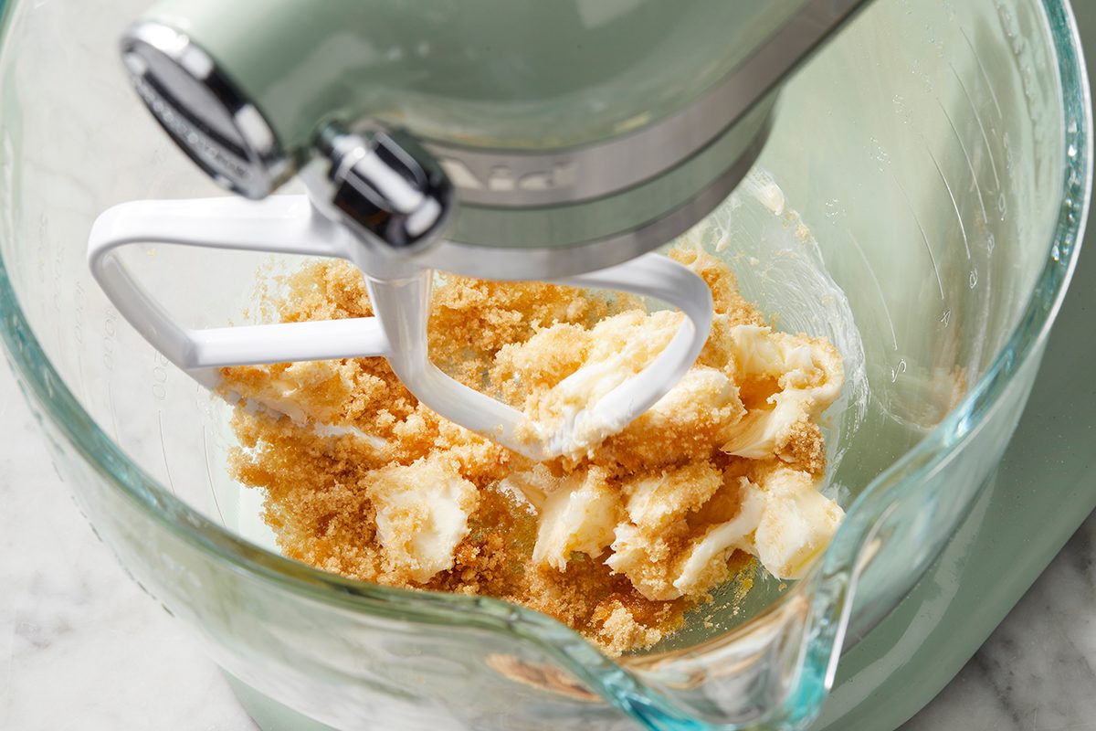 A stand mixer with a glass bowl is mixing butter and brown sugar. The ingredients are partially combined, indicating the beginning stages of a baking recipe. The mixer is set on a marble countertop.