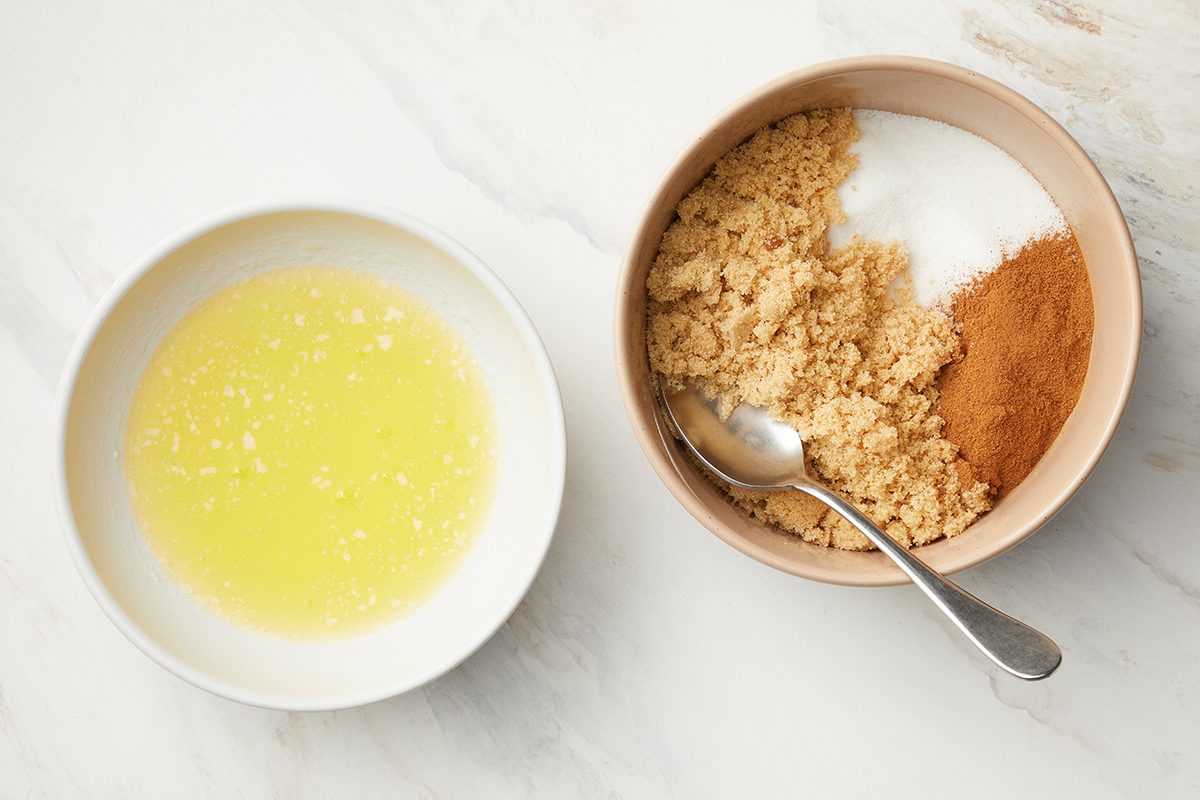 overhead shot of two bowls on a white surface, the bowl on the left contains a melted butter, the bowl on the right contains several dry ingredients, a silver spoon is placed inside the bowl with the dry ingredients, resting on top of them