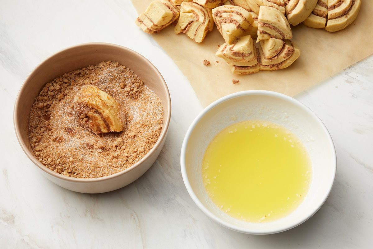 overhead shot of a kitchen counter with two bowls and parchment paper, on the left, a bowl filled with a mixture of brown crumbs and sugar holds a piece of cinnamon roll that is being dipped into the mixture, on the right, a white bowl contains melted butter, in the upper right corner, parchment paper displays several cut pieces of cinnamon rolls