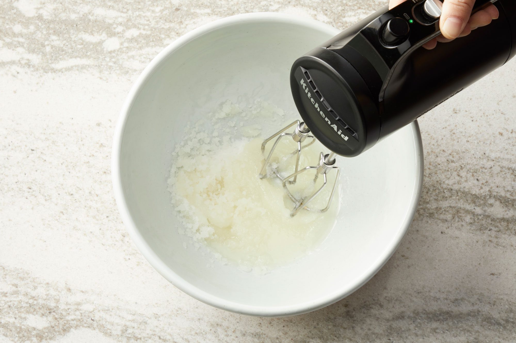overhead shot of a white mixing bowl on a light colored countertop, with the bottom of a dark colored hand mixer resting inside; the bowl contains a small amount of a light, translucent liquid, possibly egg whites, with some slightly foamy texture