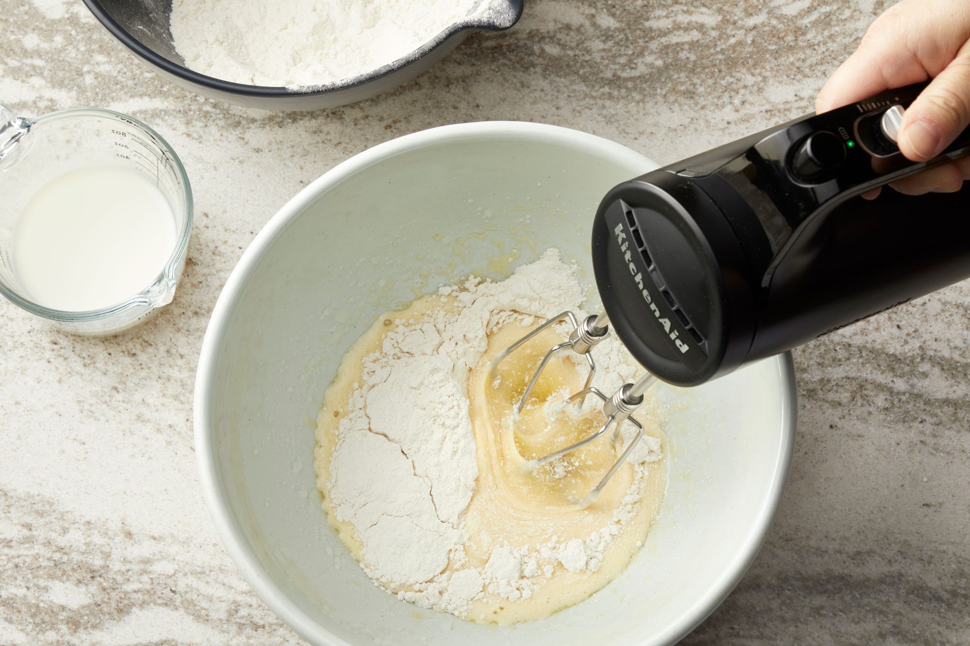 overhead shot of baking ingredients and tools on a light colored countertop, likely marble; a light green bowl sits in the center, containing a mixture of wet and dry ingredients, with flour visible on top and a yellowish batter underneath; a black KitchenAid hand mixer is positioned inside the bowl, with its beaters partially submerge in the bowl to the left, a clear glass measuring cup containing milk, sits next to the mixing bowl