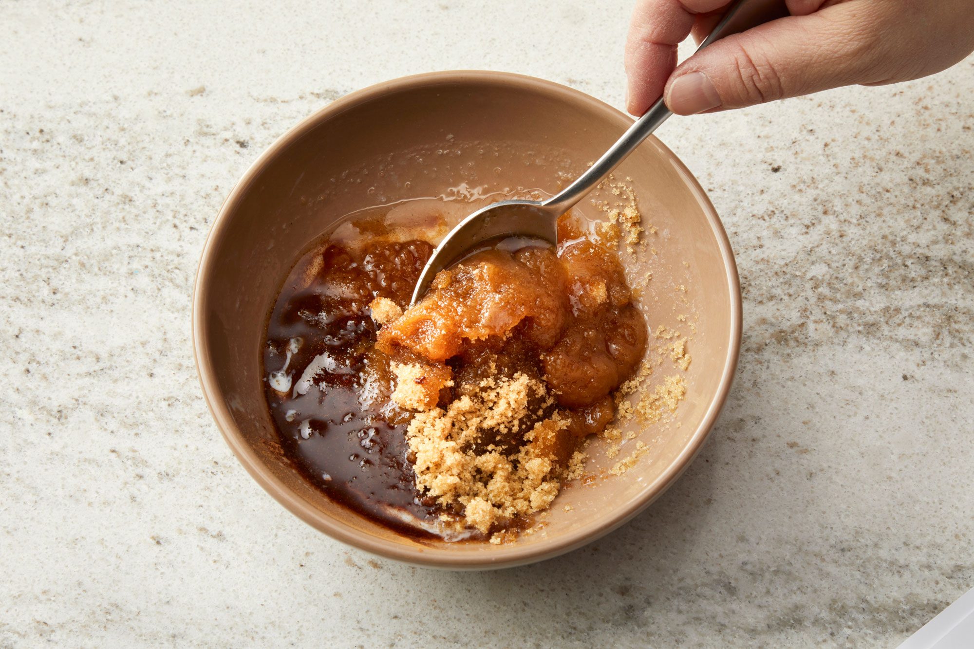 overhead shot of a tan bowl on a light colored, speckled countertop; the bowl contains a mixture of ingredients; a silver spoon rests inside the bowl, partially submerged in the mixture, with a hand visible at the top of the frame holding the spoon; the countertop appears to be a marble or granite surface