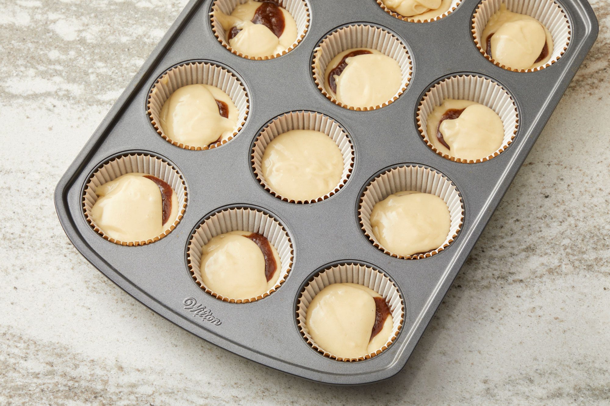 3/4th shot of a dark gray muffin tin filled with white paper liners and batter; the muffin tin is rectangular and contains twelve circular depressions; several of the liners are filled with a light colored batter, while some also contain a darker substance, likely a chocolate filling, swirled into the batter; the countertop is a light gray with a subtle speckled pattern