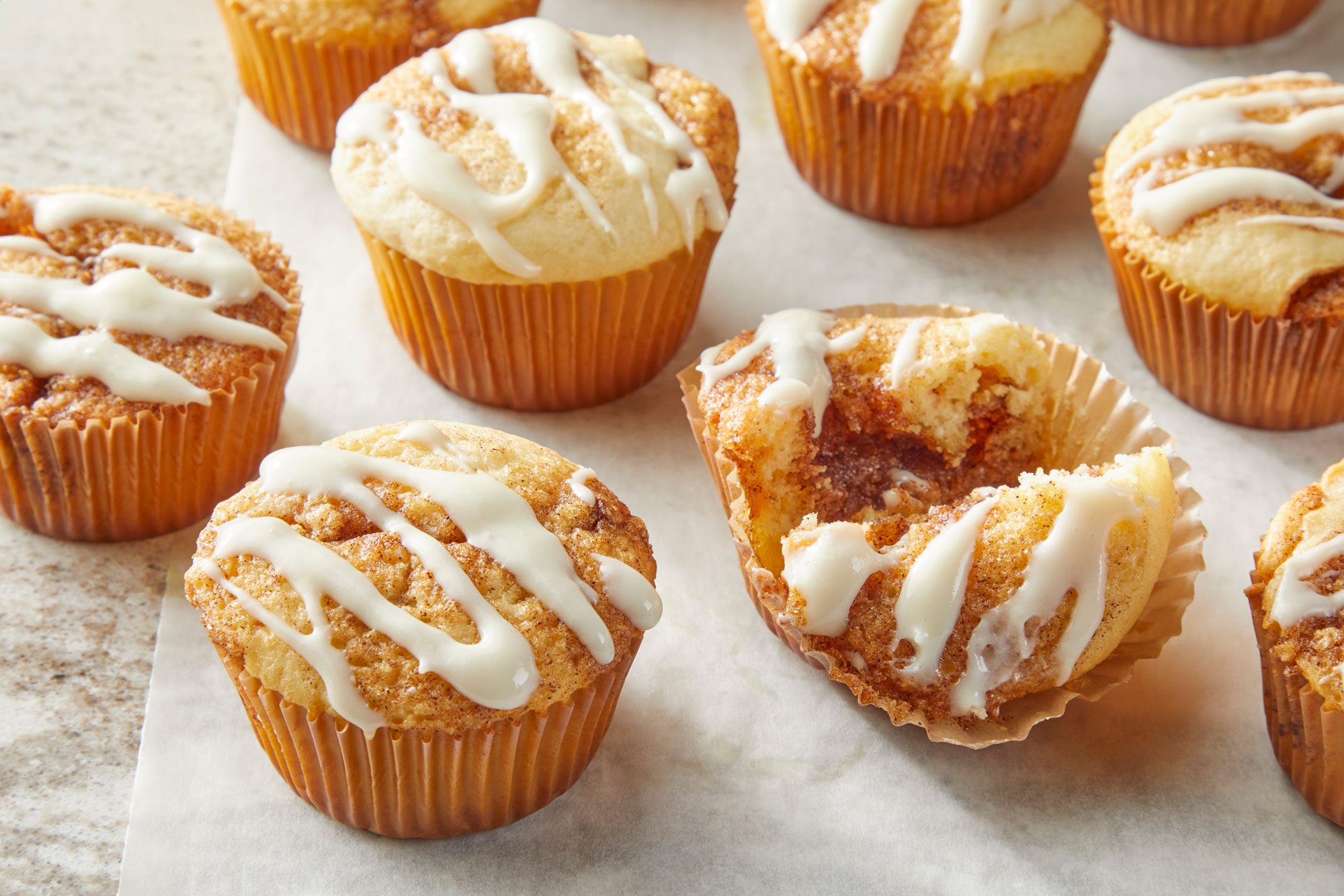 3/4th shot of several small, round baked goods on a light colored surface; and each is drizzled with a white icing in a flowing, one muffin is partially peeled open, revealing a cinnamon swirl pattern inside, the surface on which the muffins rest is a light gray