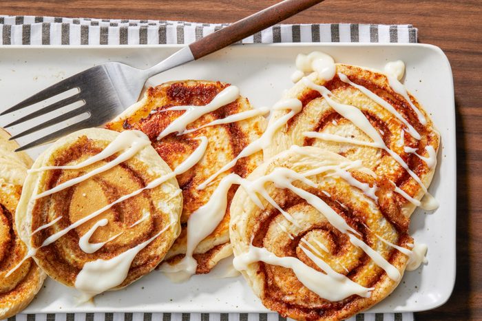 A plate of five cinnamon rolls drizzled with white icing, arranged neatly. A metal spatula rests on the plate, and the setup is on a wooden table with a striped cloth underneath.