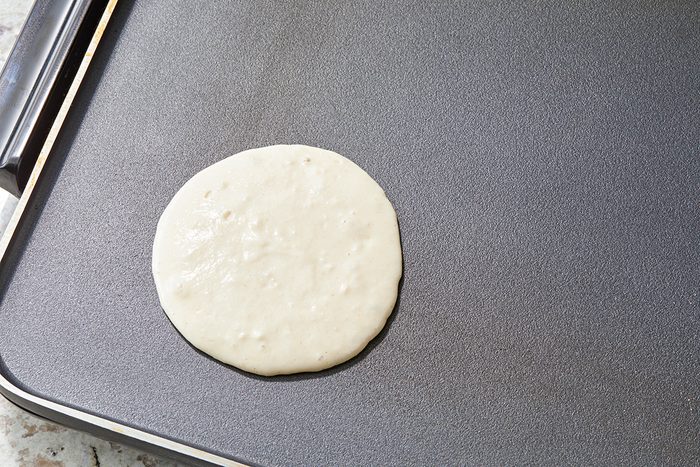 A pancake batter circle is beginning to cook on a flat, non-stick griddle surface. The background features a lightly textured countertop.