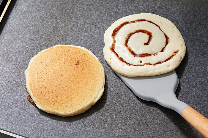 Two pancakes are cooking on a griddle. The left pancake is plain, while the right pancake has a cinnamon swirl pattern. A spatula is partially lifting the swirled pancake.