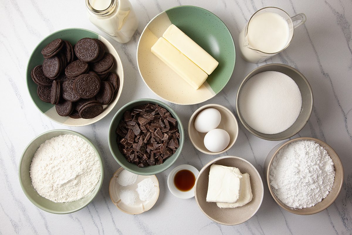 Assorted baking ingredients on a marble counter include cookies, butter, cream, sugar, flour, eggs, vanilla extract, baking powder, chocolate chunks, and cream cheese, neatly arranged in bowls and measuring cups.