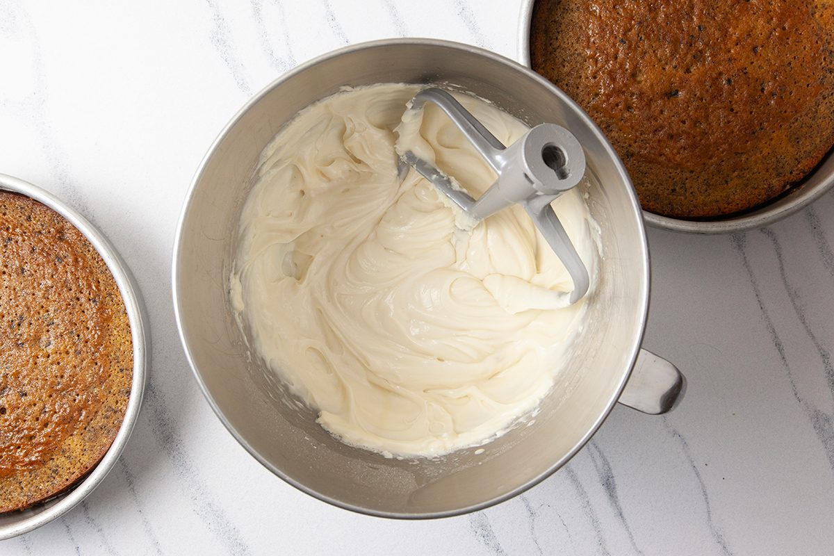 A mixing bowl of creamy white frosting with a paddle attachment, surrounded by two round cake layers on a white marble surface.
