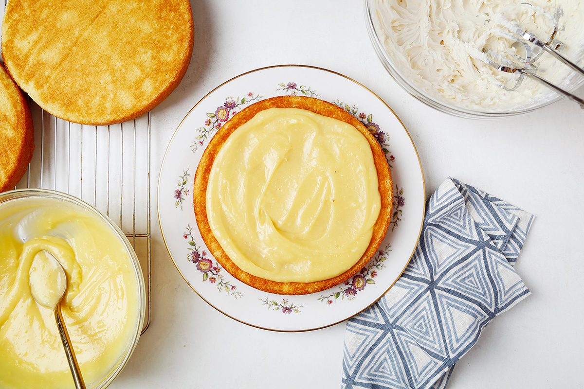 A round cake base is being frosted with yellow icing on a decorative plate. Nearby, there's a bowl of more yellow icing with a spoon, a bowl with a hand mixer, and a folded patterned cloth. Another cake layer is on a cooling rack.