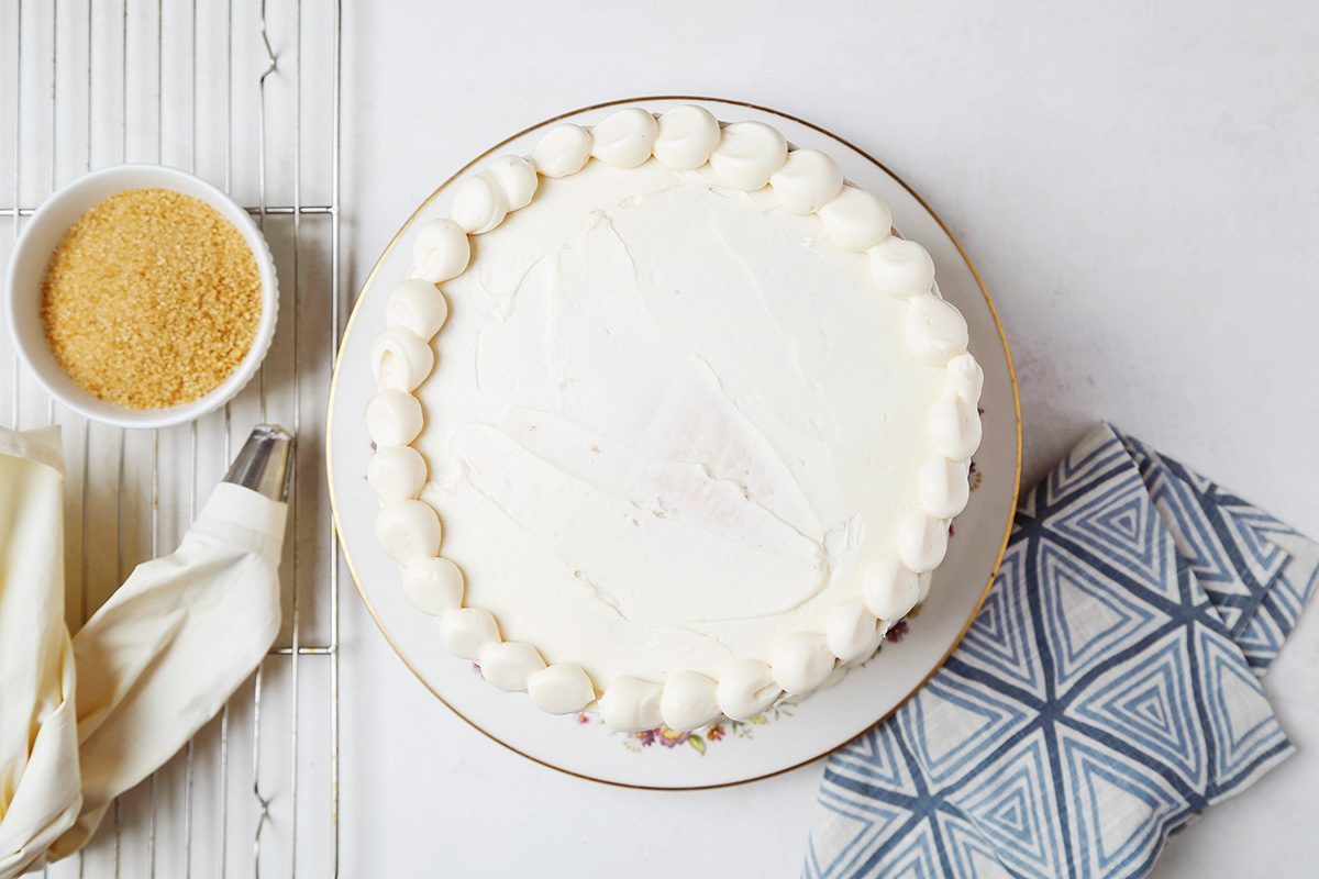 A round white cake with smooth icing and decorative piping along the edge, placed on a white plate. Nearby are a bowl of light brown sugar, a piping bag, and a blue patterned napkin on a light surface.