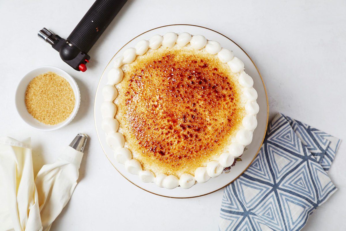 A round cake with a caramelized top, surrounding white frosting dots, and tools nearby, including a kitchen torch, piping bag, and nozzle. A small bowl of brown sugar is on the side, all set on a light surface with a patterned napkin.