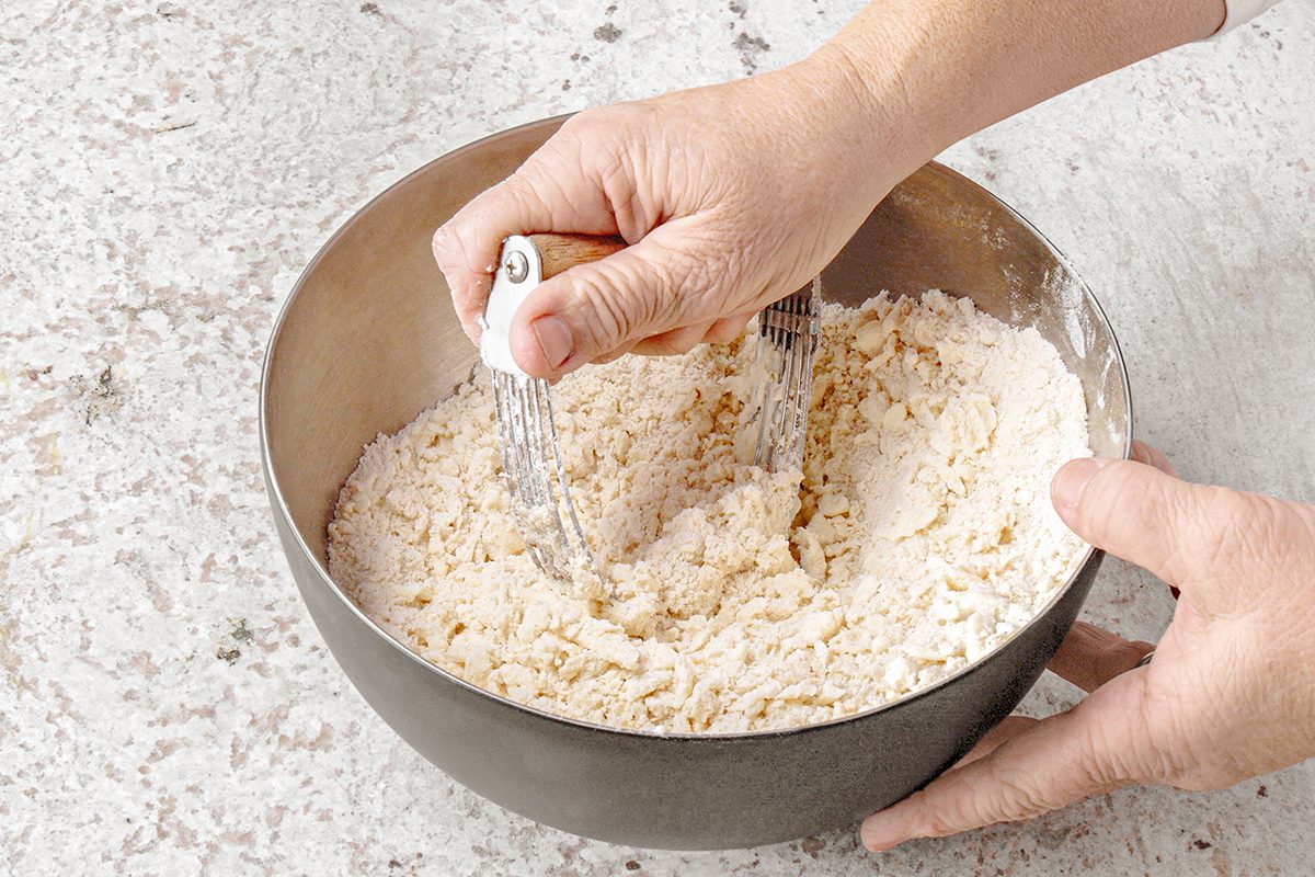 A person uses a pastry cutter to mix flour and butter in a metal bowl. The mixture appears crumbly, and the surface is light-colored with a textured countertop underneath.
