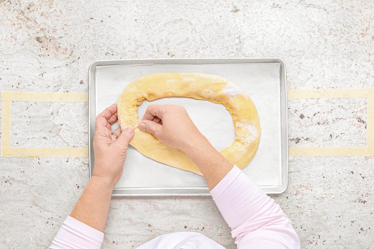 Hands shaping a piece of dough into a ring on a parchment-lined baking sheet. The background is a light marble surface.
