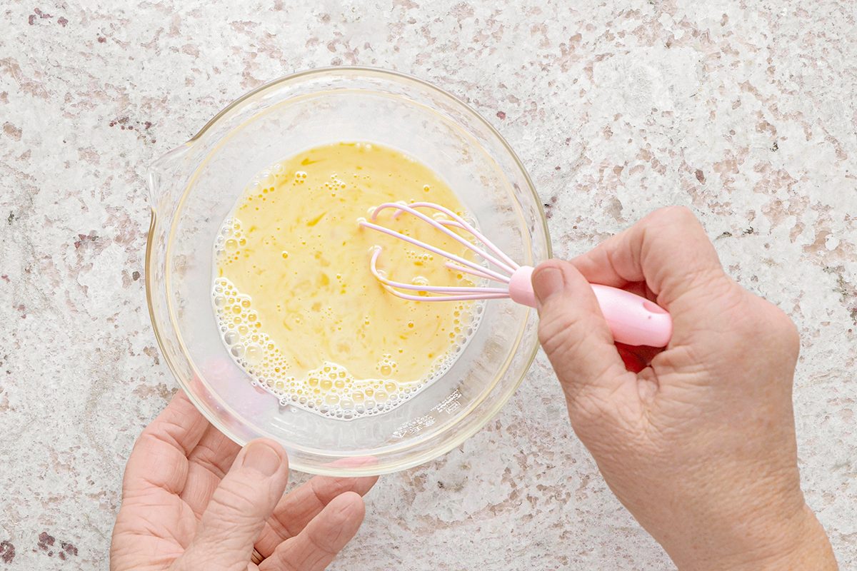Hands whisking eggs in a clear glass bowl on a speckled countertop.