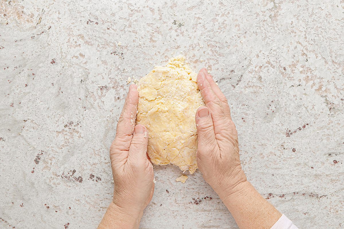 Hands shaping a piece of dough on a light-colored countertop, with scattered flour in the background.