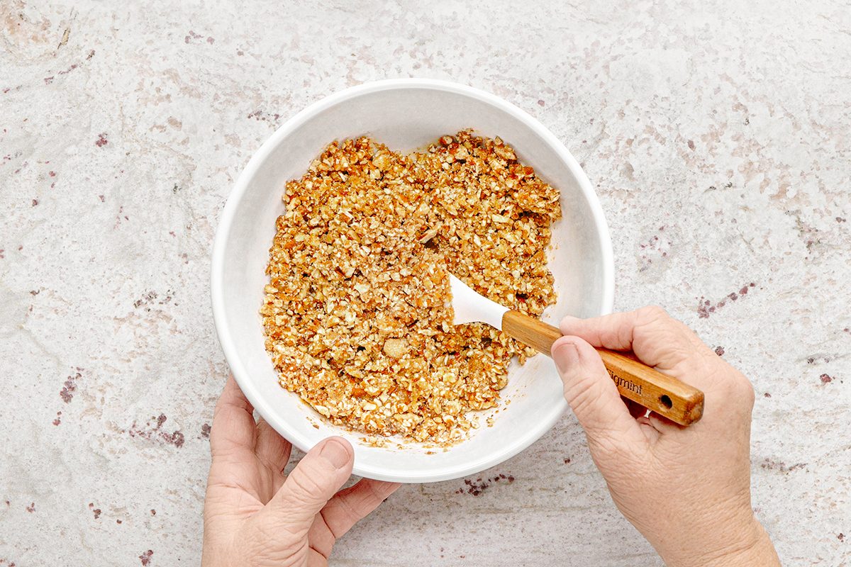 Hands mixing ingredients in a white bowl using a white spatula. The mixture appears to be a combination of chopped nuts and a caramel-like substance. The bowl is set on a speckled, light-colored countertop.