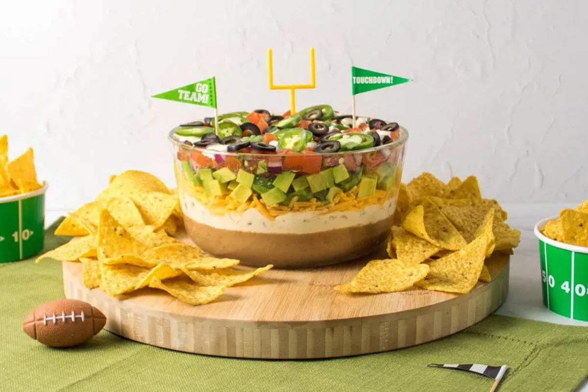7-layer dip with flags and a field goal sticking out of it, surrounded by chips and mini footballs on a wooden board and a green tablecloth