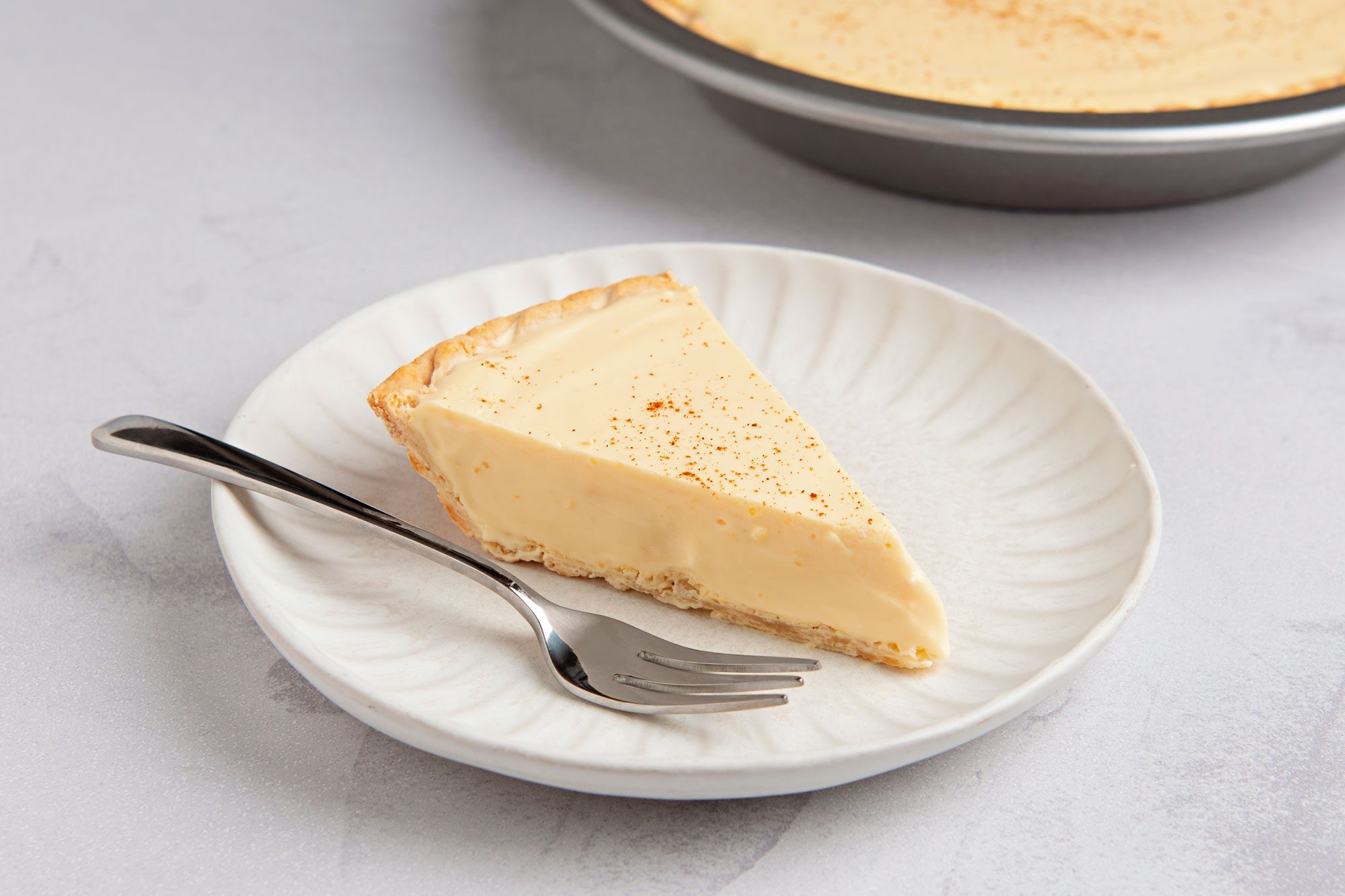 closeup shot of a slice of custard pie on a white plate with a fork next to it; the pie is dusted with cinnamon
