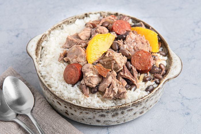A bowl containing white rice topped with chunks of meat, sliced sausage, black beans, and orange segments.
