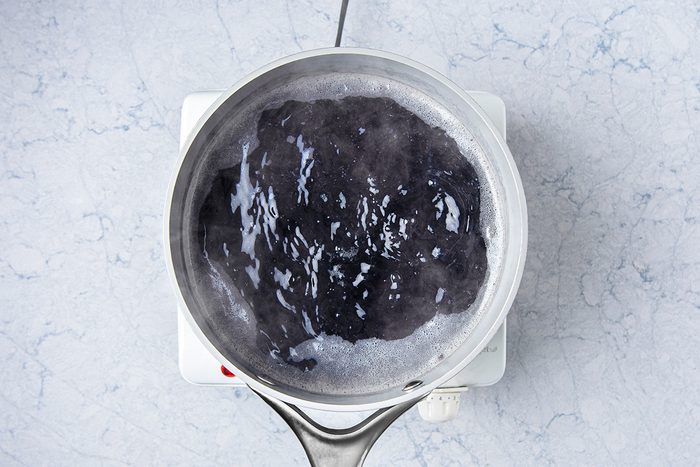 A stainless steel pot with boiling water on a white electric stovetop, viewed from above. The water appears dark, possibly infused with a dark ingredient, creating a rippling, bubbling effect over a light marbled surface.