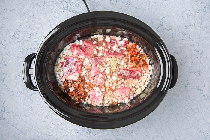 Overhead view of a slow cooker with uncooked ingredients, including chunks of raw meat, chopped onions, and spices mixed with broth. The slow cooker sits on a marble countertop.