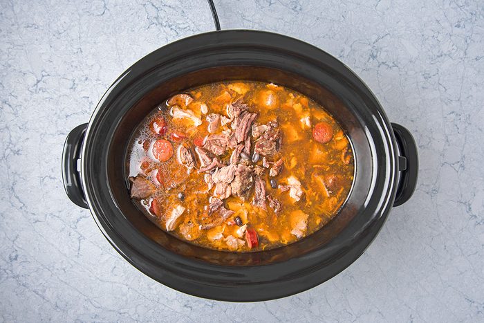 A black slow cooker filled with a hearty beef stew. The stew includes chunks of beef, sliced carrots, mushrooms, and a rich, red-brown broth.