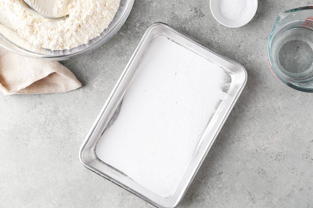 A rectangular baking pan with a white powdery layer inside sits on a gray surface. Nearby are a bowl of flour, a small bowl with white powder, and a clear glass measuring cup. A beige cloth is partially visible next to the flour bowl.