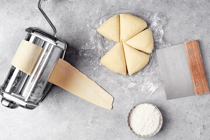 A pasta maker with dough being rolled out, beside a circle of dough divided into six triangles. A bowl of flour and a metal dough scraper are also on the gray countertop.