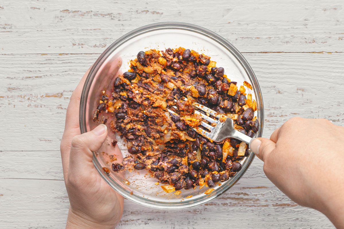 overhead shot of a person mixing black beans with a fork in a glass bowl; the bowl is sitting on top of a white wooden table
