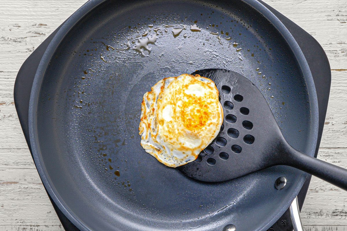 overhead shot of a single fried egg in a non-stick pan, the egg is cooked on one side and still in the pan; it is being flipped by a black spatula with holes; the pan is sitting on a white wooden surface