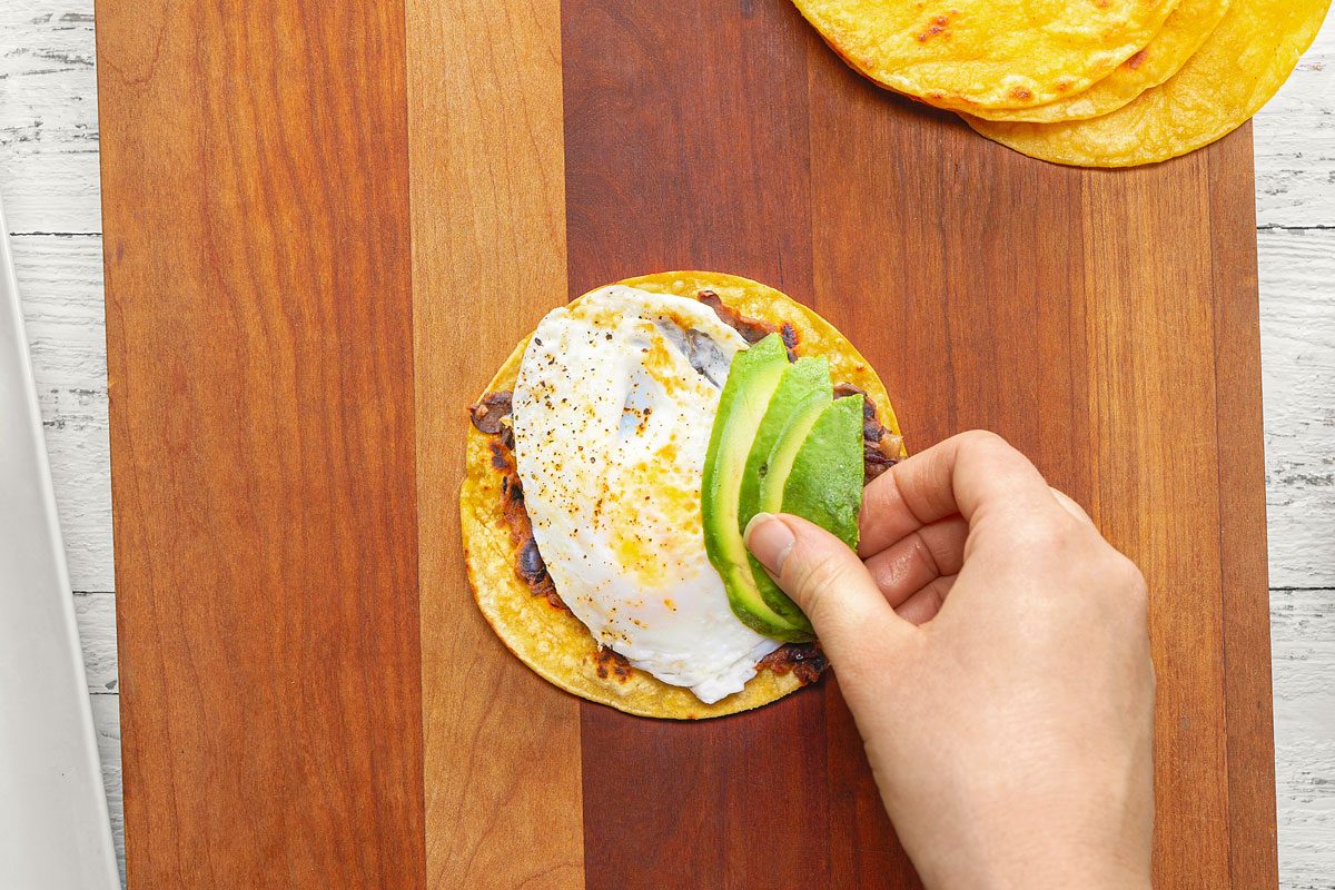 a person making a breakfast taco on a wooden cutting board; the taco is made with a corn tortilla, a fried egg, refried beans, and avocado slices; there is a plate of fried eggs in the background