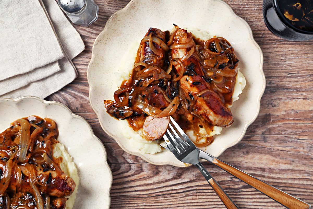 Irish Bangers and Mash shot on a brown wooden surface. Bangers and gravy served on top of mashed potatoes on scalloped plates; overhead